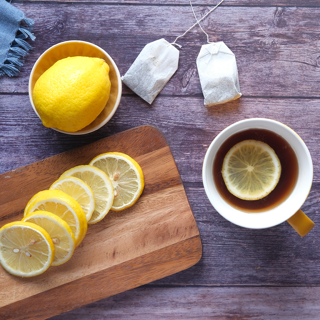 Cup of tea with lemon slice on a wooden table, below two tea bags and next to a cutting board with lemon slices. Above the cutting board is a bowl with a lemon.