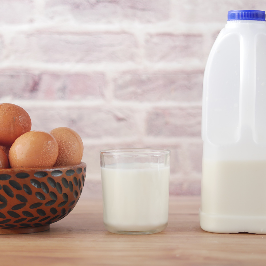 Glass of milk on a wooden table in front of a brick wall. On the left are eggs in a bowl and on the right is a half gallon of milk.