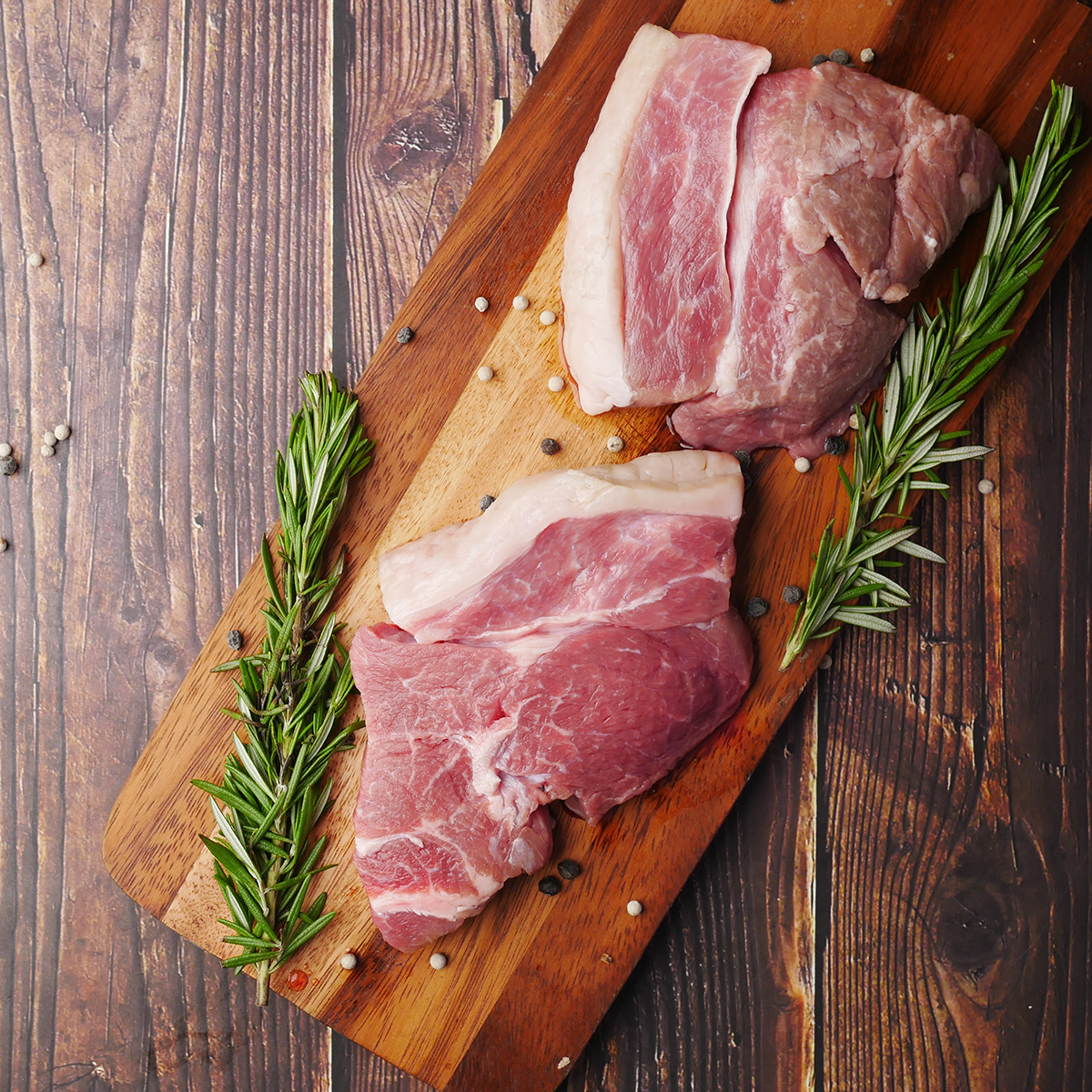 Two raw meats on brown wooden chopping board with green garnish and spices on top of a wooden table.