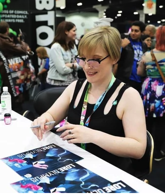 Rachel Smythe signing autographs at a convention. She has lanyards around her neck, glasses on, blond hair and is wearing a black tank top.